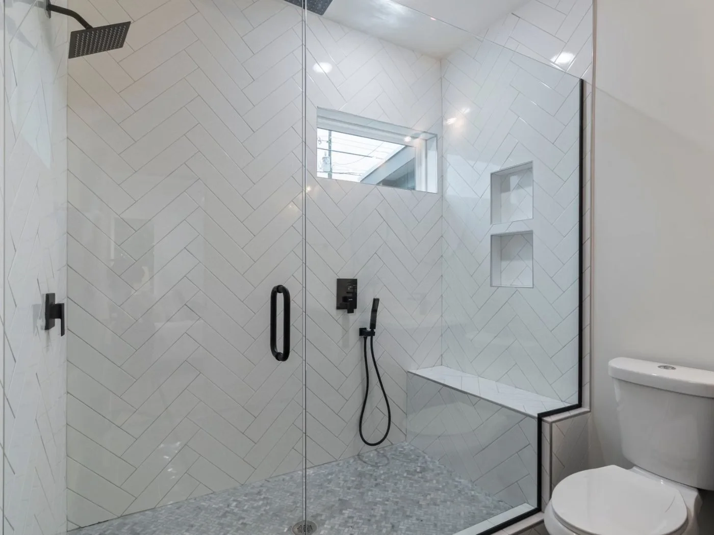 Accessible bathroom remodel Longview WA â€” a walk-in shower with a clear glass door, a handheld shower on a vertical slide bar, a built-in corner seat and a recessed niche in herringbone marble tile, with a toilet alongside