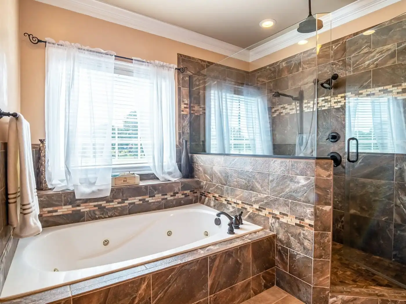 Bathroom remodel Castle Rock WA — a drop-in whirlpool tub set under a curtained window beside a glass walk-in shower, in brown marble-effect tile with a mosaic accent band and oil-rubbed bronze fittings