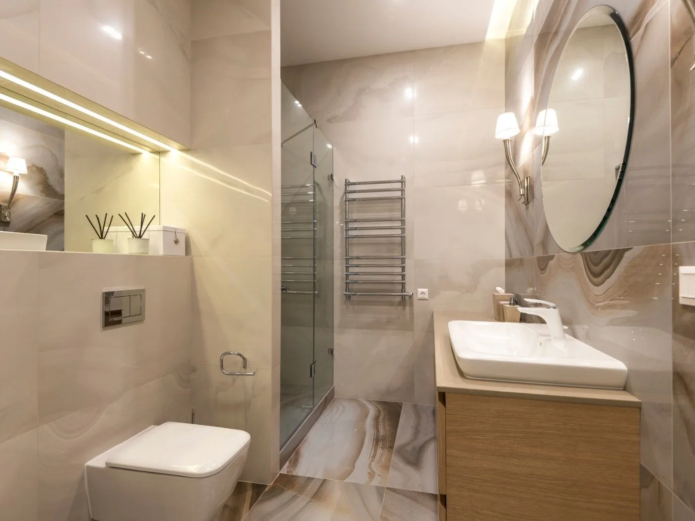 Bathroom remodel Kalama WA — a warm stone-tiled bathroom with a round mirror over a wood vanity and basin, a heated towel rail and a glass shower screen beyond