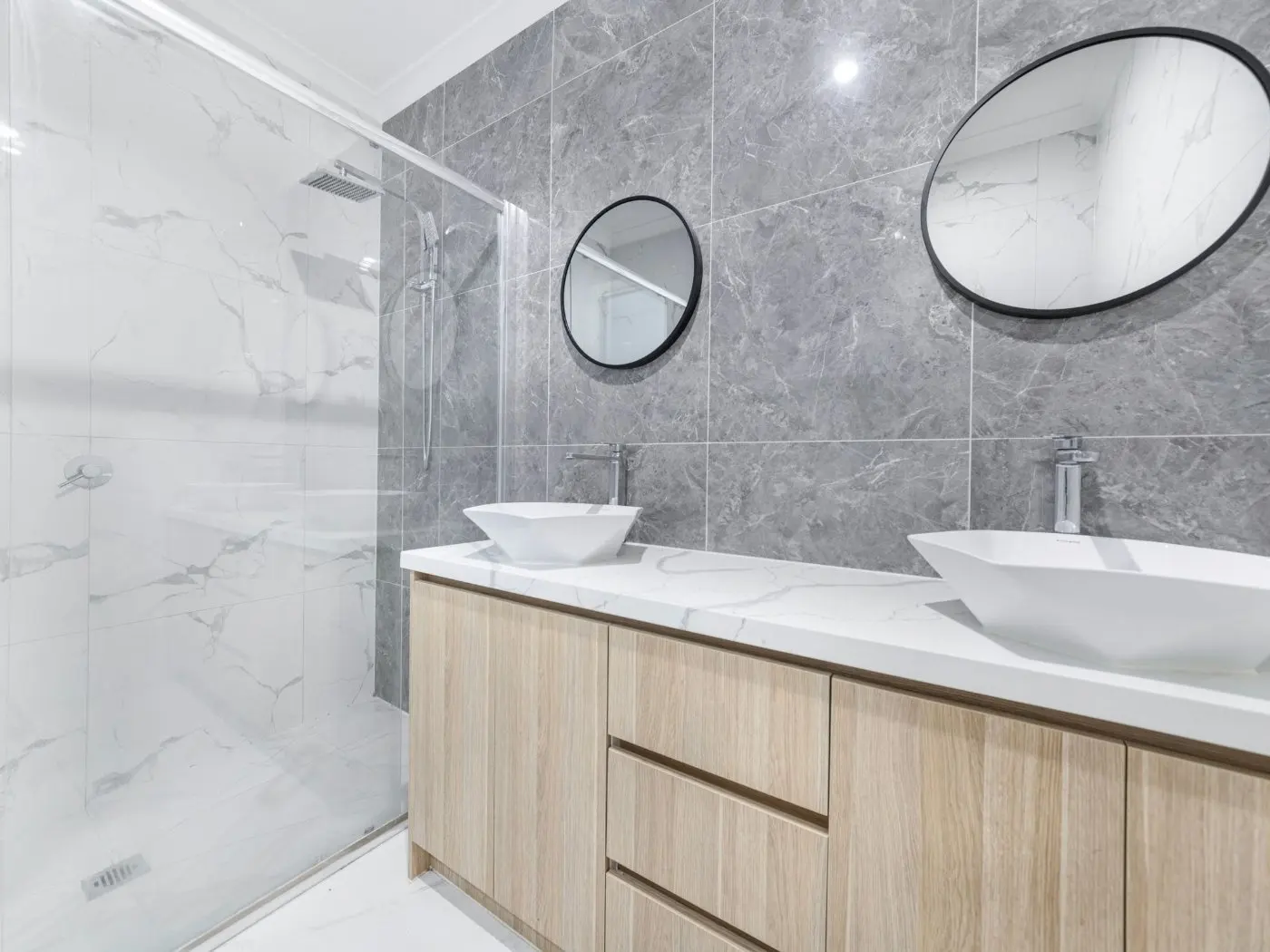 Bathroom remodel West Longview — two white vessel basins on a light oak floating vanity, round black-framed mirrors and a glass walk-in shower with a rainfall head, in grey marble-effect tile