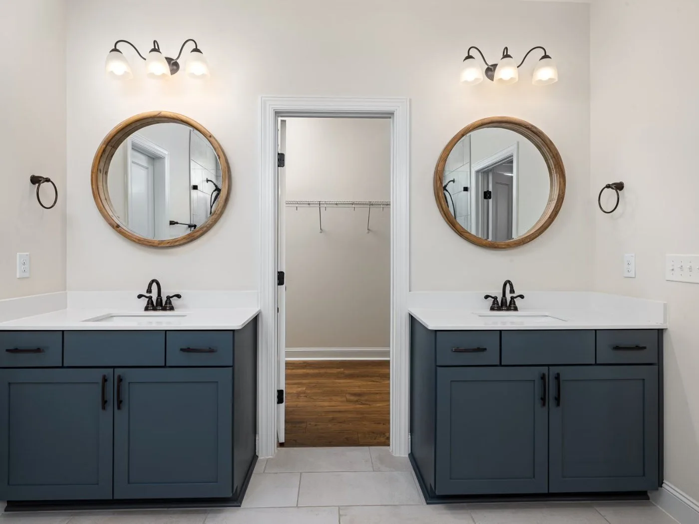 Bathroom vanity installation Longview WA â€” two navy blue shaker vanities with white countertops, undermount basins and oil-rubbed bronze three-hole faucets, each below a round wood-framed mirror and a three-light bar fitting