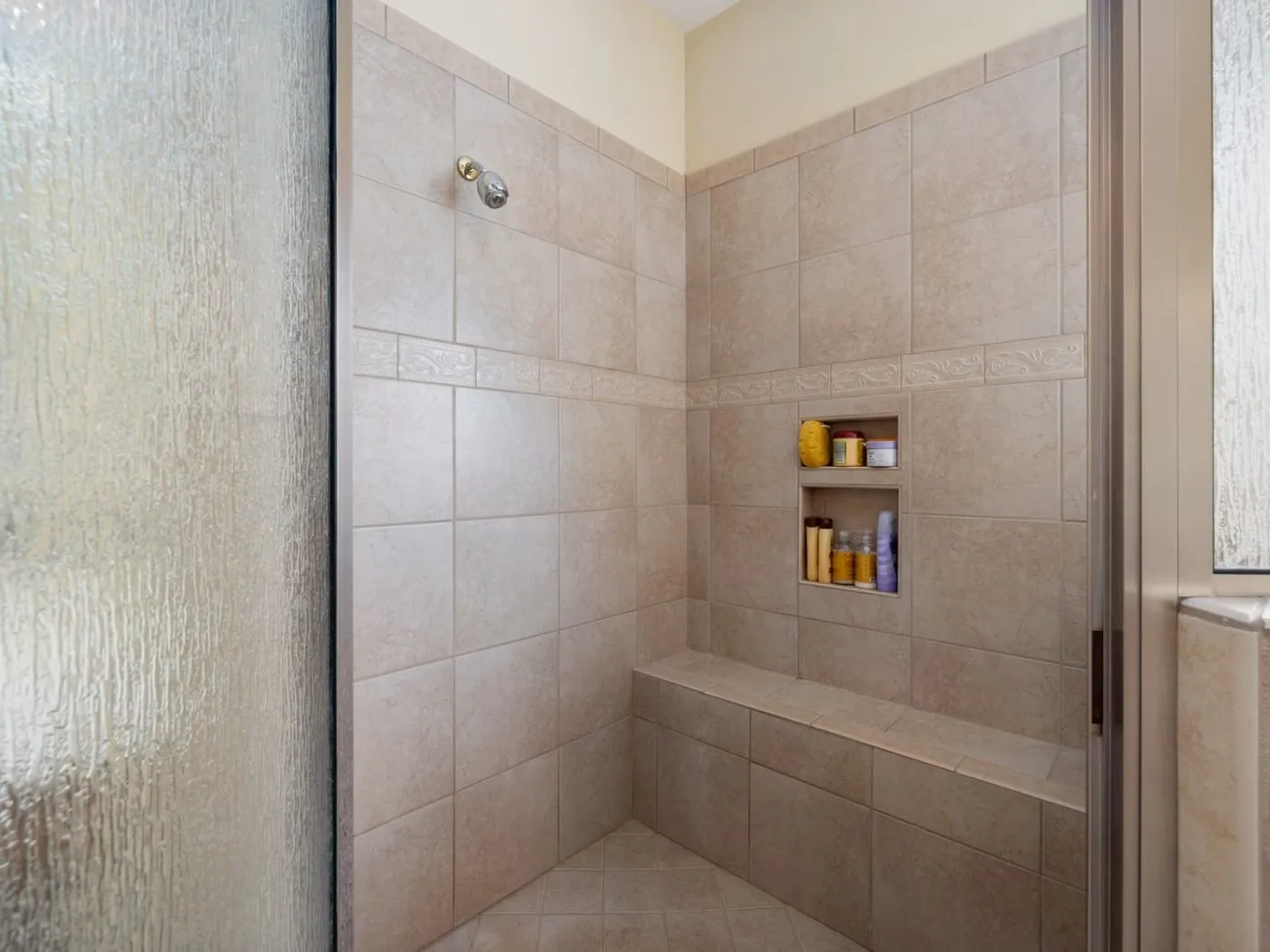 Walk-in shower installation Longview WA — a beige stone-tiled walk-in shower with a glass panel, a built-in bench and recessed shelving where a bath used to sit
